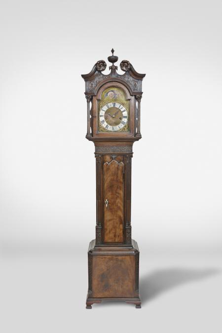 England, miniature tall clock, 8 day, time and strike on bell, weight driven movement in a carved Chinese Chippendale style mahogany case with broken arch top, carved rosettes, center finial, and plinth and foliate carving below molding on the cornice on hood, the dial flanked by two full stop fluted columns with carved capitals above trunk with shaped door and inlaid escutcheon flanked by stop fluted quarter columns with carved capitals resting on a molded base with carved and chamfered corners on ogee style feet. The brass dial has silvered chapter ring, painted moon disc and cast spandrels, signed in center "William Wilks, London"