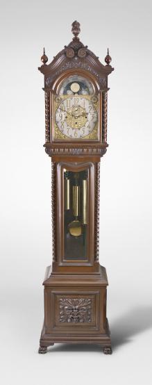 Tall clock, 8 day weight driven movement, time, strike and chime on gongs, in an elaborately carved mahogany case with broken arch top surmounted by three carved finials including center flaming urn, twist columns flanking hood and trunk doors, both of which have beveled glasses, fretwork sides on hood, over a base with carved Northwind center panel and resting on paw feet. The brass dial has silvered chapter ring, applied brass numerals, moon phase, seconds bit, and strike / silent and tune selection discs.