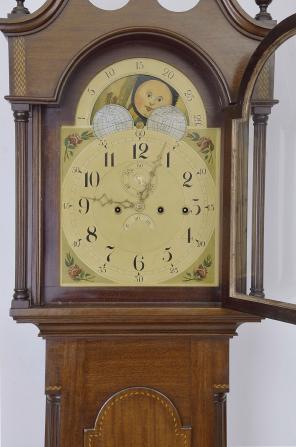 Tall clock, probably English, 8 day, time, strike weight driven movement with Westminster chime on auxiliary movement, in an inlaid mahogany case with broken arch top, three wooden finials, full fluted columns flanking hood over tombstone trunk door with string and shell inlay, flanked by fluted quarter columns resting on a base with string and shell inlay and flared French feet. The unsigned painted metal dial has moon phase, date calendar and painted rose spandrels.