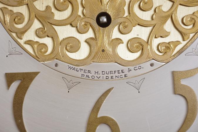 Walter Durfee & Co., Providence, Rhode Island, hall clock, 8 day, time, strike and chime on nine tubular bells playing choice of Whitington or Westminster chimes, three brass weight driven movement by Elite with a Winterhalder & Hofmeier trademark. The mahogany case has arched top with two brass finials, surmounted by carved crest over a carved keystone, applied carvings, full fluted columns flanking hood door, and fretted sides. The trunk has carved egg and dart molding below hood, with beveled glass and carved top, flanked by two full Corinthian columns, all resting on paw feet. The brass dial has silvered chapter ring, subsidiary seconds, chime / silent and tune selector discs with moon phase indicator, engraved brass spandrels, applied brass numerals, and fretted center, and is stamped "ELITE" with trademark and "GERMANY" on lower edge of dial.