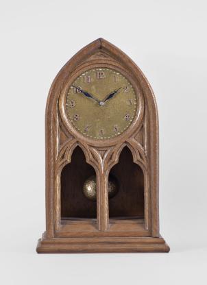 National Magnetic Clock Co. (Tiffany), Buffalo, NY, "Model 2001" electro-mechanical shelf clock, battery-powered, time only movement in a gothic form oak case with a pair of trefoil squint window apertures to view the pendulum, hammered brass dial with applied metal numerals, and two-ball torsion pendulum, serial number 5001.