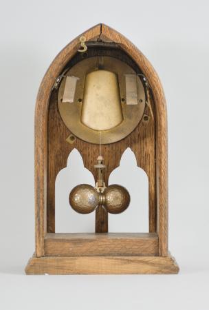 National Magnetic Clock Co. (Tiffany), Buffalo, NY, "Model 2001" electro-mechanical shelf clock, battery-powered, time only movement in a gothic form oak case with a pair of trefoil squint window apertures to view the pendulum, hammered brass dial with applied metal numerals, and two-ball torsion pendulum, serial number 5001.