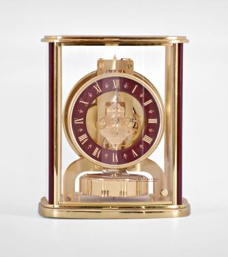 Jaeger-LeCoultre, Switzerland, Atmos perpetual clock, 528 caliber, jeweled movement with torsion pendulum in a polished brass case with curved acrylic panels, round maroon enamel dial with incised roman numerals, and polished brass hands.