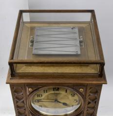 J.B. Winter, Brighton, England, a standing, rolling ball clock of unique design, the front of the square, pedestal form oak case with simple, carved gothic inspired ornament below and flanking the signed, arabic numeral metal dial, and top with molded edge, glazed, oak framed cover for the steel tray with channels for guiding the rolling ball, the case with removable sides and back to access the movements, the interior of the case filled with various machinery including the table movement with electromagnetic release for tilting the tray and bevel gear with clutch to advance the dial movement, electric motor at bottom with centrifugal governor, with bevel gearing and vertical shaft with clutch used to periodically rewind the spring that tilts the table, with one steel and two brass balls