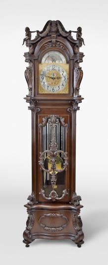 Herschede Electric Clock Co., Cincinnati, Ohio, a monumental hall clock, sounding Westminster, Whittington or Canterbury quarters on nine tubular bells, electrically rewound, spring driven time train, pendulum regulated movement in a highly carved mahogany case, with incurved serpentine base on scroll feet, carved pseudo pilasters flanking the glazed trunk door with stylized anthemion, acanthus and bowknot suspended fruit, the bonnet with carved console brackets flanking a break arch door, acanthus carving, and molded arch with stylized keystone, surmounted by a scroll pediment with central carved shell, festoons, and carved finials of unclothed children, silvered and brass dial with moon phase and tune selector