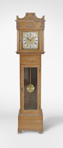 Self Winding Clock Co., New York, a striking, quarter sawn oak hall clock with Neoclassical ornament, squat rectangular base on bracket feet and ogee molding with applied composition acanthus leaf ornament, the trunk with rectangular glazed door, beaded molding, composition urn, and composition egg and dart molding, the bonnet with full turned columns, the sides with stained glass panels and simple Arts and Crafts inspired fret, flanking a square door and supporting an entablature with Grecian ornament including full palmette leaves and scrolling vine, ogee molded cornice with applied acanthus ornament, surmounted by a gallery with reeded pilasters and crest with flower filled cornucopia and trailing ribbon, composite dial with silvered roman numeral chapter ring, matted center, subsidiary silvered seconds chapter, and gilt spandrels, fancy pierced blued steel hands, Seth Thomas movement with Self Winding