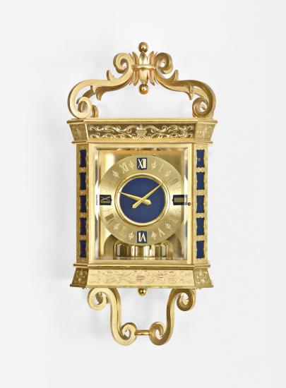 Jaeger LeCoultre, for Gubelin, Switzerland, an unusual hanging Atmos timepiece, the gilt case with four engraved scroll ornaments at base and top, each joining a gilt ring at the center, and the top ring with stylized crown, canted corners with engraved, pierced blind fret revealing a blue painted ground, canted, engraved base and cornice, the front and sides with thick beveled glasses, roman numeral gilt dial with blue, shield shaped cartouches at 3, 6, 9, and 12:00, and slightly sunk blue center, gilt hands and typical mid 20th century jeweled Atmos movement with lever escapement, and with gilt, T shaped mounting bracket