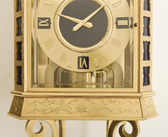 Jaeger LeCoultre, for Gubelin, Switzerland, an unusual hanging Atmos timepiece, the gilt case with four engraved scroll ornaments at base and top, each joining a gilt ring at the center, and the top ring with stylized crown, canted corners with engraved, pierced blind fret revealing a blue painted ground, canted, engraved base and cornice, the front and sides with thick beveled glasses, roman numeral gilt dial with blue, shield shaped cartouches at 3, 6, 9, and 12:00, and slightly sunk blue center, gilt hands and typical mid 20th century jeweled Atmos movement with lever escapement, and with gilt, T shaped mounting bracket