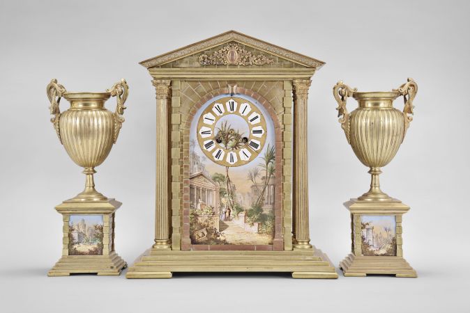 France, three piece mantel garniture set, 8 day, time and strike, spring driven pendule de Paris movement in a metal case of classic architectural form with stepped base, six full Corinthian columns flanking an ashlar facade with central arch, a painted ceramic panel within displaying a gilt chapter ring with Roman numerals, a scene of royals walking amidst an ancient city below the dial, all topped by a pediment with applied gilt ornament, two matching urns, both with painted ceramic panels.