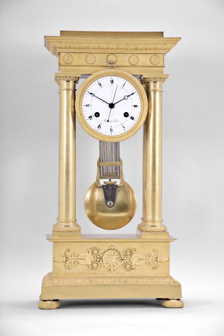 Boileau a Paris, an early 19th century, half seconds beating gilt bronze portico clock, on floral medallion ornamented compressed ball feet, base molding decorated with palmette, anthemion and bellflower, the dado with mount having a central sunflower with mask, and flanking Grecian foliate ornament below the simple molded surbase, four columns with square plinths, laurel wreathed molded bases, and egg and dart capitals supporting the entablature, the frieze with floral medallions and cornice with waterleaf, palmette and bellflower, laurel wreath bezel with simple foliate inner ring, roman numeral white enamel dial with inner seconds chapter,signed "Boileau a Paris", blued steel Breguet style hands and central seconds hand, large two train movement with knife edge suspension and pin wheel escapement, counting the hours on a bell, and heavy bimetallic half seconds beating gridiron pendulum