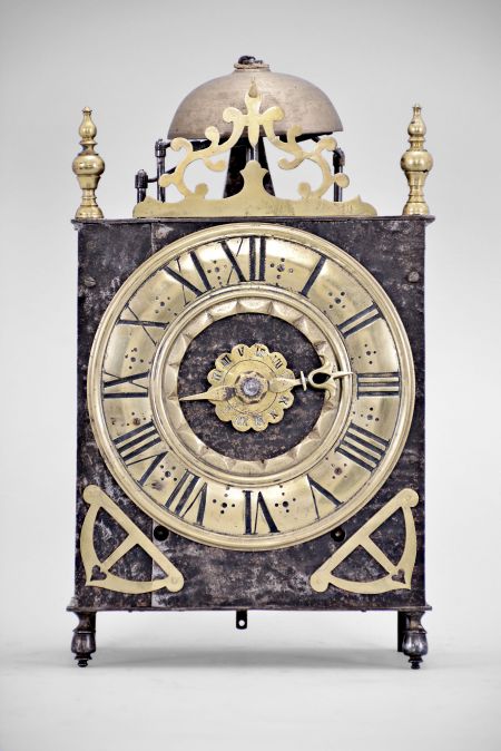 France, Comtoise lantern clock, 8 day, time and strike with alarm, weight driven movement with verge escapement, thread suspension with rear-hanging pendulum in a posted iron frame case, heavy brass chapter ring with Roman numerals, single brass hand, alarm ring with Roman numerals, applied brass spandrels with hearts, all resting on turned iron feet in the front, and topped with a thick pierced brass fronton flanked by two turned brass finials.