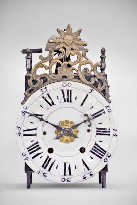 France, Comtoise lantern clock, 8 day, time and strike with alarm, weight driven movement with verge escapement, thread suspension with rear-hanging pendulum in an iron post and plate frame, early faïence dial with raised portions under each Roman numeral, outer ring with Arabic numerals every five minutes, two steel hands, brass alarm ring with Arabic numerals, topped by a cast brass fronton with image of the sun / Apollo flanked by two turned iron finials, all standing on tapered iron feet.