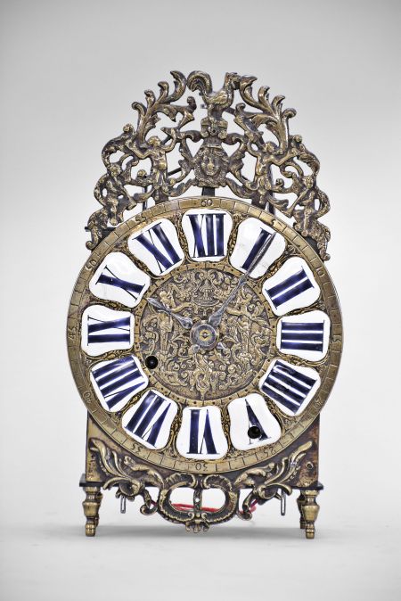 France, Comtoise lantern clock, 8 day, time and strike, weight driven condensed movement with verge escapement, wire suspension with rear-hanging pendulum in an iron posted case with hammered brass doors, cast brass dial with inset white enamel cartouches with blue Roman numerals, brass outer ring with Arabic numerals every 5 minutes, two steel hands, topped by cast bronze fronton featuring trumpeters and a forward facing Gallic rooster, all sitting on four turned brass feet.