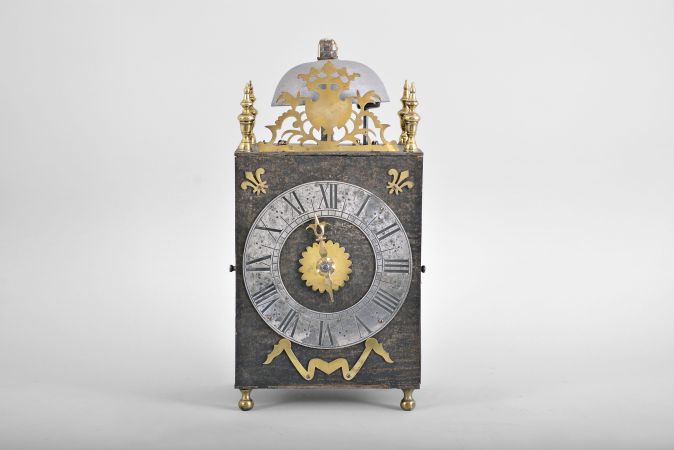 France, Comtoise lantern clock, 8 day, time and strike, weight driven movement with verge escapement, thread suspension with rear-hanging pendulum in a iron post and plate frame, pewter chapter ring with engraved Roman numerals, single brass hand, applied brass fleur de lis spandrels and pivoted winding arbor shutters, all resting on four turned brass feet, and topped by flat pierced brass fronton flanked by four turned brass finials.