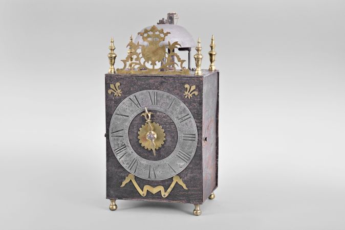 France, Comtoise lantern clock, 8 day, time and strike, weight driven movement with verge escapement, thread suspension with rear-hanging pendulum in a iron post and plate frame, pewter chapter ring with engraved Roman numerals, single brass hand, applied brass fleur de lis spandrels and pivoted winding arbor shutters, all resting on four turned brass feet, and topped by flat pierced brass fronton flanked by four turned brass finials.