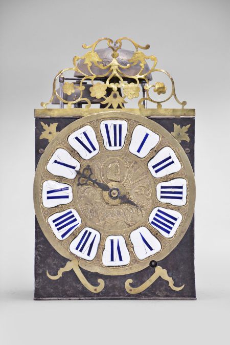 France, Comtoise clock, 8 day, time and strike with alarm, weight driven movement with rare Maillet (or Mayet) escapement and unusual double vertical rack strike in a posted iron frame, cast brass dial with profile of young Louis XV with inset white enamel cartouches with blue Roman numerals, alarm ring with Arabic numerals, single pierced iron hand, brass spandrels above and pivoted winding arbor shutters below, all topped with a pierced brass fronton with floral motif.