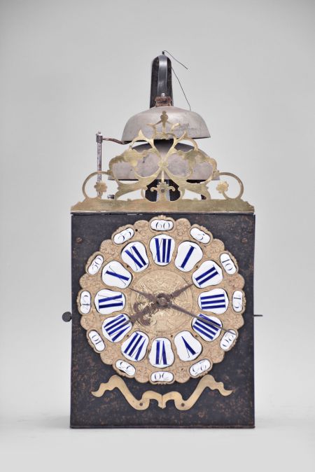 France, Comtoise clock, 8 day, time and strike with two bells, weight driven movement, verge escapement, thread suspension with rear-hanging pendulum in an iron post and plate frame, cast brass dial with two iron hands, floral motif and inset with 24 white enamel cartouche numerals, blue Roman numerals for the hours and black Arabic numerals every five minutes, flat brass winding arbor shutters below the dial, and pierced brass fronton.