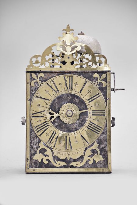 Brocard, Comté, France, Comtoise clock, 8 day, time and alarm, weight driven movement with verge escapement, thread suspension with rear-hanging pendulum in a iron post and plate frame, brass chapter ring with engraved Roman numerals, brass alarm ring with engraved Arabic numerals, flat brass fleur de lis spandrels and pivoted winding arbor shutters, engraved plaque below the dial reading "Brocard en Comté", single pierced brass hand, all topped with a pierced brass fronton.