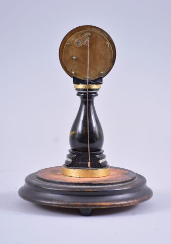 A small, European candlestand clock, ebonized baluster form base with floral decoration supporting a roman numeral white enamel dial with blued steel Breguet style hands, 30 hour timepiece movement with tic tac escapement, all mounted to a decorated turned wooden base with glass dome