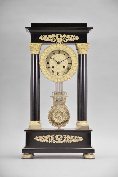 France, Empire portico clock, 8 day, time and strike, spring driven pendule de Paris movement in a polished black slate case on cast brass feet, the base and top with applied gilt rose ornaments, four Corinthian columns with gilt bases and capitals, the gilt bezel with repeating acanthus leaf design, gilt chapter ring with Roman numerals, cast dial center and gridiron pendulum both with floral designs, blued steel Breguet hands.