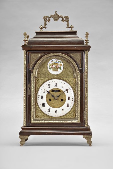 Austria, a late 18th century bracket clock, the molded base with cast, gilt feet, faux arch door with fabric backed frets at the upper corners, and caddy top, surrounded by four finials and with carrying handle, engraved brass dial with roman numeral, white enamel chapter ring, surrounded by cast spandrels, blued steel hands, patterned center with aperture for observation of mock pendulum, and boss in the arch with polychrome enamel scene with bird and basket of flowers, two train, two day movement with crown wheel escapement and rack striking, sounding the hours on a bell